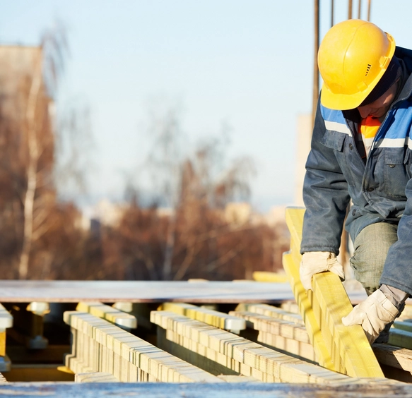 Un artisan charpentier avec un casque de sécurité jaune ajuste des poutres en bois sur une structure de toit en construction. Le travail précis et minutieux assure une charpente solide et durable