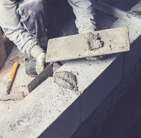 Un artisan maçon avec truelle à ciment, vêtu de vêtements de travail, applique soigneusement du mortier sur une structure en construction à Saint-Julien-les-Rosiers