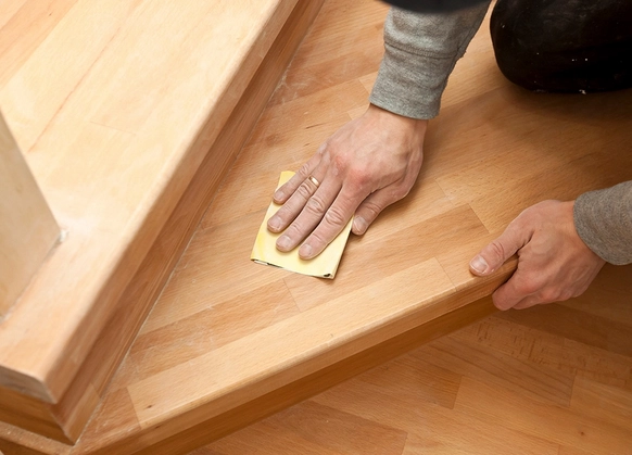 Artisan ponçant un escalier en bois à l'aide de papier de verre pour une finition lisse et soignée. Ses mains montrent un travail minutieux, garantissant une surface impeccable