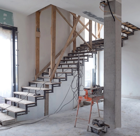 Escalier en construction avec structure métallique et marches en bois, entouré d'échafaudages. L'intérieur est en cours de rénovation, avec des outils et des câbles visibles sur le chantier